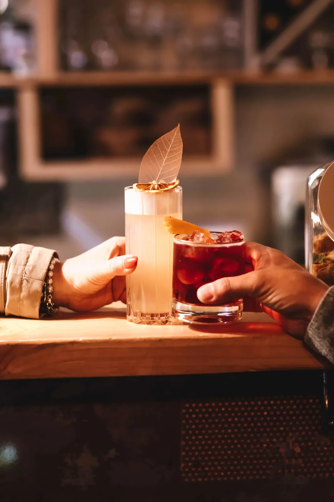 Due mani si scambiano due cocktail sul bancone, uno con foglia e arancia essiccata